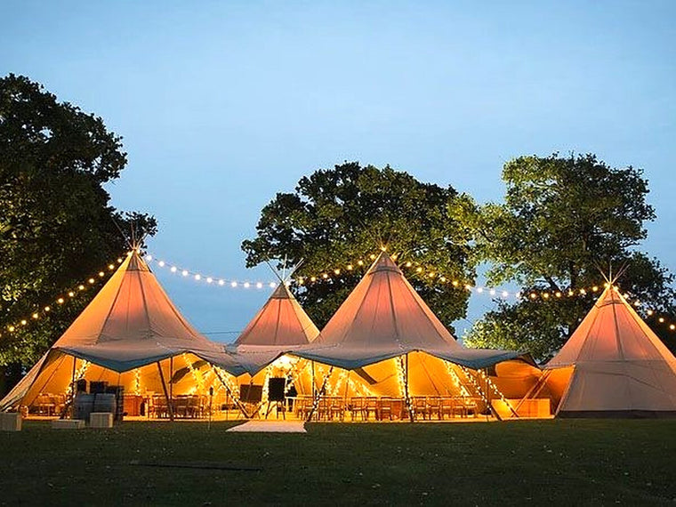 Four linked wedding tipis illuminated at dusk beneath festoon lights.