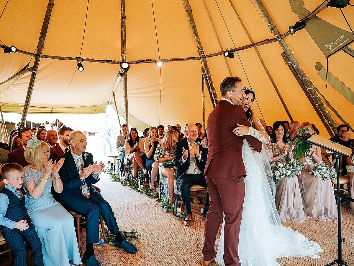 Newlyweds embrace as guests applaud inside a tipi ceremony.